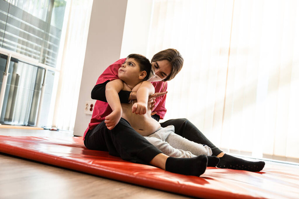 инвалид ребенок и физиотерапевт на красном гимнастическом коврике делает упражнения. пандемическая защита маски - Фото, изображение