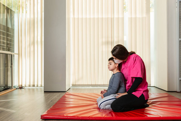 mozgássérült gyermek és fizioterapeuta egy piros tornatermi szőnyegen gyakorlatokat végez. pandémiás maszkvédelem - Fotó, kép
