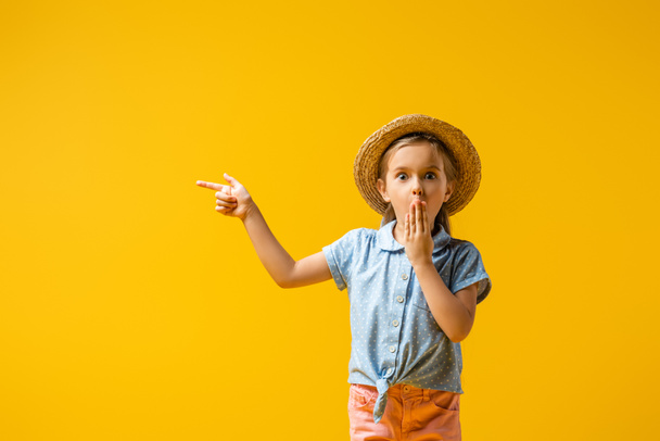 шокированный ребенок в соломенной шляпе, прикрывающий рот и указывающий пальцем, изолированным на желтый - Фото, изображение