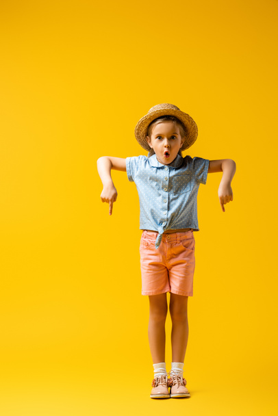 pełna długość wstrząśniętego dziecka w słomkowym kapeluszu skierowanym w dół palcem na żółto - Zdjęcie, obraz
