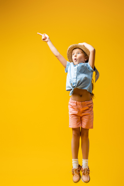 longitud completa de niño sorprendido en sombrero de paja apuntando con el dedo en amarillo - Foto, imagen