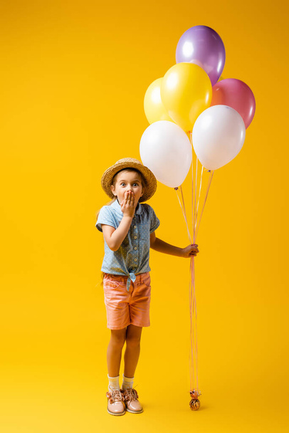 full length of shocked kid in straw hat covering mouth and holding balloons on yellow - Photo, Image