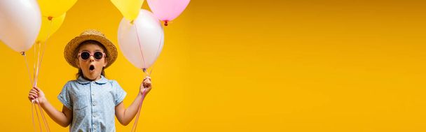 шокированный ребенок в соломенной шляпе и солнцезащитных очках держа воздушные шары изолированы на желтом, баннер - Фото, изображение
