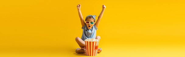 ενθουσιασμένοι παιδί σε 3d γυαλιά κάθεται με σταυρωμένα πόδια και σήκωσε τα χέρια κοντά σε κουβά ποπ κορν και βλέποντας ταινία σε κίτρινο, πανό - Φωτογραφία, εικόνα
