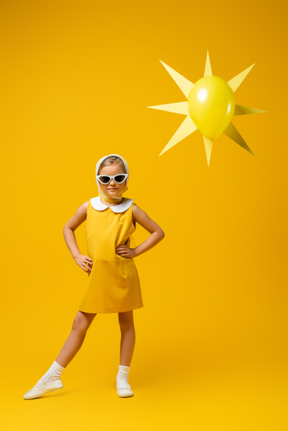 piena lunghezza della ragazza in velo e occhiali da sole in piedi con le mani sui fianchi vicino al sole decorativo con palloncino su giallo - Foto, immagini