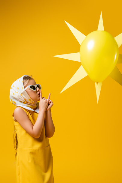 tyttö huivi ja aurinkolasit osoittaa hiljainen merkki lähellä koriste aurinko ilmapallo eristetty keltainen - Valokuva, kuva