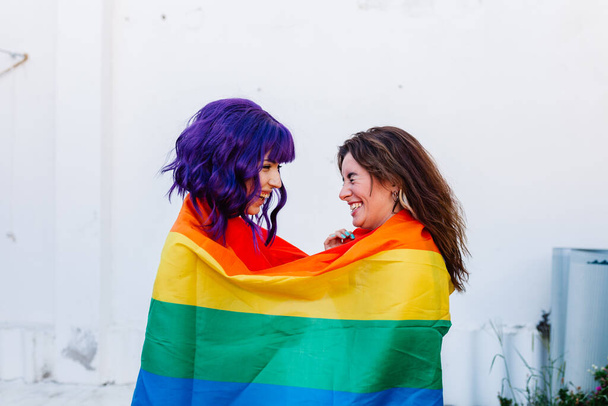 Zwei Frauen mit Regenbogenfahne auf einem Hinterhof. Junge lesbische Mädchen halten eine Regenbogenfahne in der Hand und umarmen sich glücklich. Ein Paar lesbische Frauen - Foto, Bild