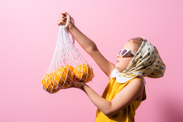 girl in headscarf and sunglasses holding reusable string bag with oranges isolated on pink - Photo, Image