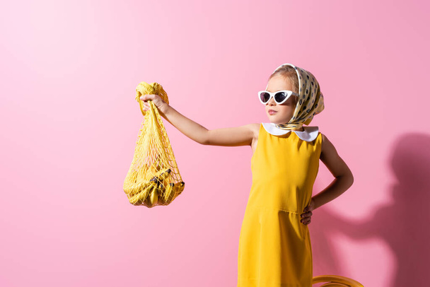 menina em lenço de cabeça e óculos de sol segurando saco de corda reutilizável com bananas, enquanto de pé com a mão no quadril em rosa - Foto, Imagem
