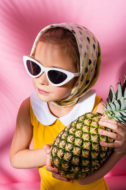 Kind mit Kopftuch und Sonnenbrille hält Ananas auf rosa  - Foto, Bild
