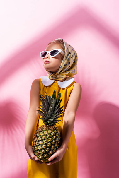 stylish girl in headscarf and sunglasses holding pineapple on pink  - Photo, Image