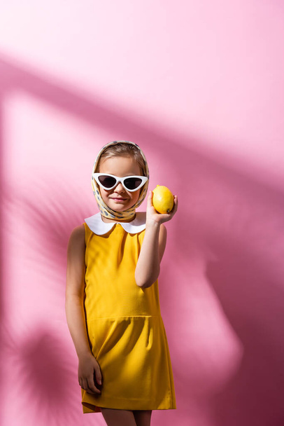 счастливая девушка в платке и солнечных очках с лимоном на розовом  - Фото, изображение