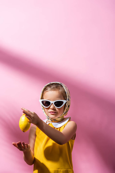 stylish girl in headscarf and sunglasses holding whole lemon on pink  - Zdjęcie, obraz