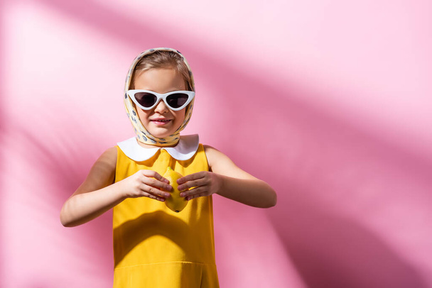 улыбающаяся девушка в платке и солнцезащитных очках, держащая свежий лимон на розовом  - Фото, изображение