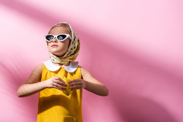 stilvolles Mädchen mit Kopftuch und Sonnenbrille mit frischer Zitrone auf rosa  - Foto, Bild