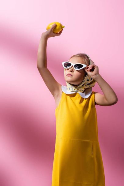 elegante niño en pañuelo para la cabeza y gafas de sol con limón por encima de la cabeza en rosa  - Foto, Imagen