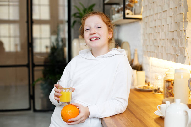 Rapariga com cabelo encaracolado vermelho bebe suco de laranja em pé na cozinha, comida saudável para o bebê. - Foto, Imagem