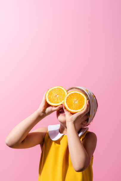 wesoła dziewczyna pokrywa oczy trzymając pomarańczowe połówki odizolowane na różowy  - Zdjęcie, obraz