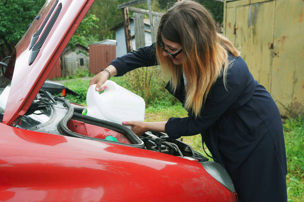 Eine charmante junge Frau steht in der Nähe der offenen Motorhaube eines Autowracks, versucht es zu reparieren, gießt Flüssigkeit ab - Foto, Bild