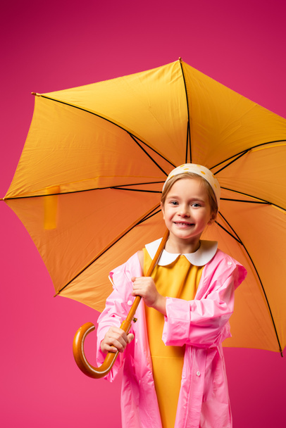 pleased girl in raincoat standing under umbrella isolated on crimson - Fotoğraf, Görsel