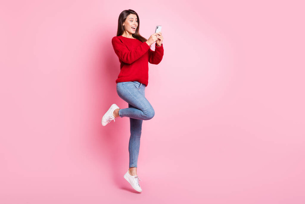 Full length photo of cute girl jump excited screen hold telephone wear red sweater denim jeans sneakers isolated pink color background - Foto, Imagem