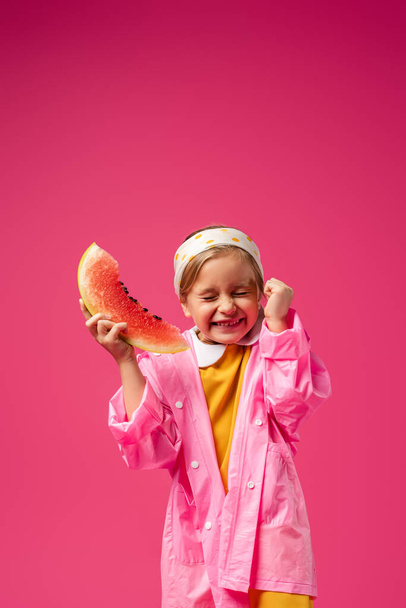 opgewonden meisje in regenjas houden watermeloen op karmozijn - Foto, afbeelding