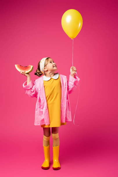 longitud completa de chica alegre en impermeable sosteniendo globo y sandía en rodajas en carmesí - Foto, Imagen