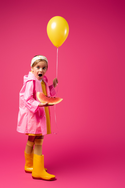 volle Länge des erstaunten Mädchens im Regenmantel mit Luftballon und Wassermelone in Scheiben auf Karminrot - Foto, Bild