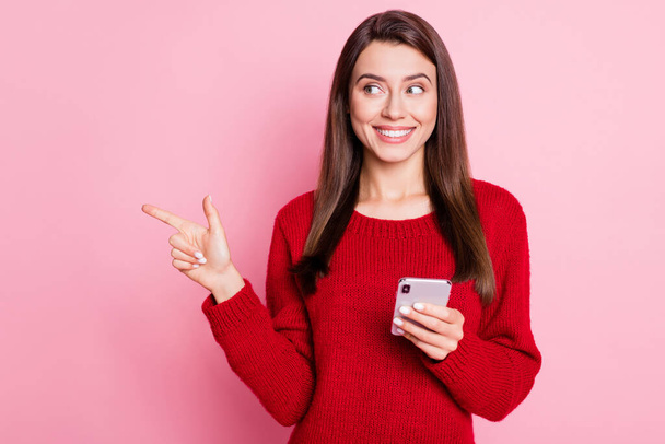 Фото прекрасной девушки, направляющей указательный палец смотреть сторону пустое пространство держать сотовый телефон блестящая улыбка носить красный свитер изолированный розовый цвет фона - Фото, изображение