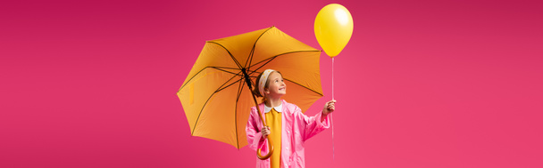 χαρούμενο κορίτσι με αδιάβροχο κρατώντας μπαλόνι και κίτρινη ομπρέλα απομονωμένη σε πορφυρό, banner - Φωτογραφία, εικόνα