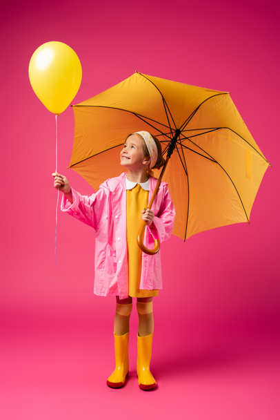 full length of cheerful girl in raincoat holding balloon and yellow umbrella on crimson - Photo, Image