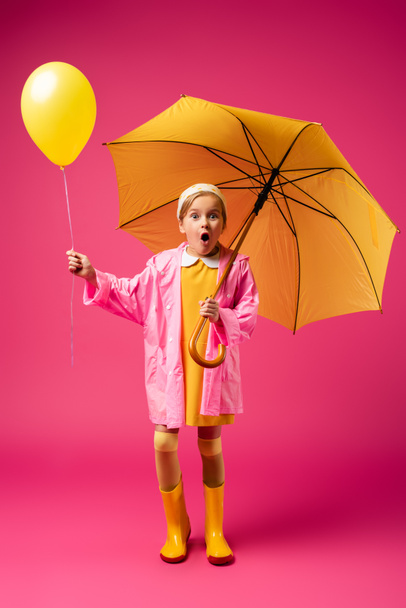 full length of amazed child in raincoat and rubber boots holding balloon and yellow umbrella on crimson - Φωτογραφία, εικόνα