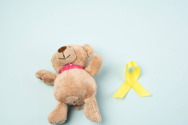brown teddy bear holds in his paw a yellow ribbon folded in a loop on a blue background. concept of the fight against childhood cancer. problem of suicides and their prevention - Foto, imagen