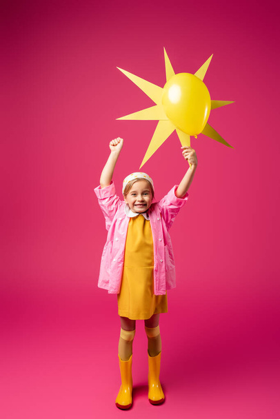 full length of happy girl in raincoat and rubber boots holding decorative sun with balloon on crimson - Photo, Image