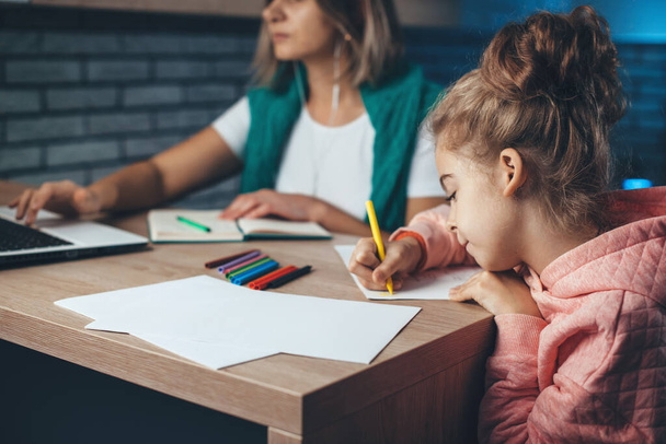 Caucasian mother and is working on a laptop with business stuff while her daughter is drawing with colorful pencils - Φωτογραφία, εικόνα