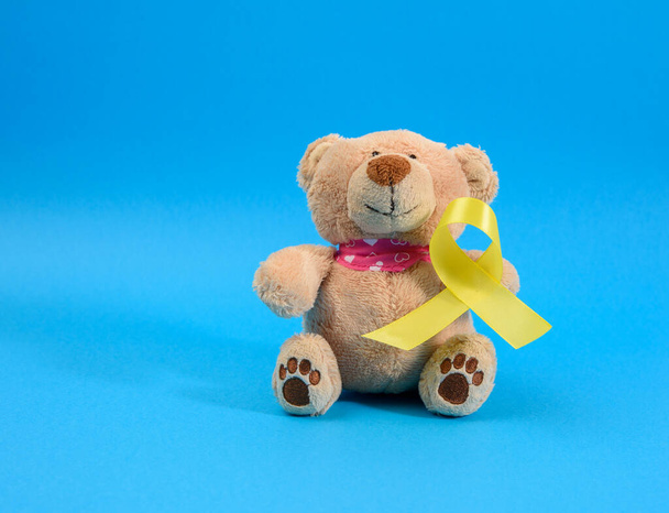 brown teddy bear holds in his paw a yellow ribbon folded in a loop on a blue background. concept of the fight against childhood cancer. problem of suicides and their prevention - Photo, Image