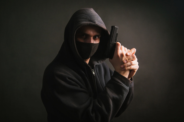 Een gangster in een capuchon, een masker, houdt een wapen in zijn handen op een donkere achtergrond. De bandiet mikt met een pistool. Foto in de studio. - Foto, afbeelding