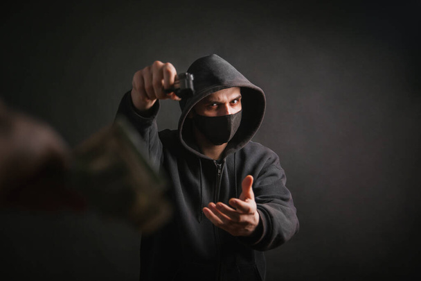 Ein Gangster mit Kapuze und Maske hält vor dunklem Hintergrund eine Waffe in der Hand und nimmt dem Opfer Geld ab. Der Räuber zielt mit seiner Pistole und fordert Dollars. - Foto, Bild