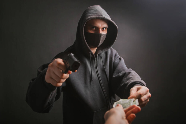 A gangster in a hood and a mask holds a weapon in his hands on a dark background and takes money from the victim. The bandit aims his pistol and demands dollars. - 写真・画像