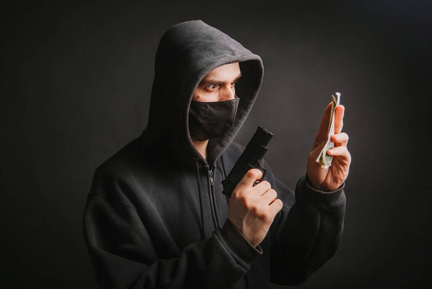 A gangster in a hood and a mask holds a weapon in his hands on a dark background and takes money from the victim. The bandit aims his pistol and demands dollars. - Foto, imagen