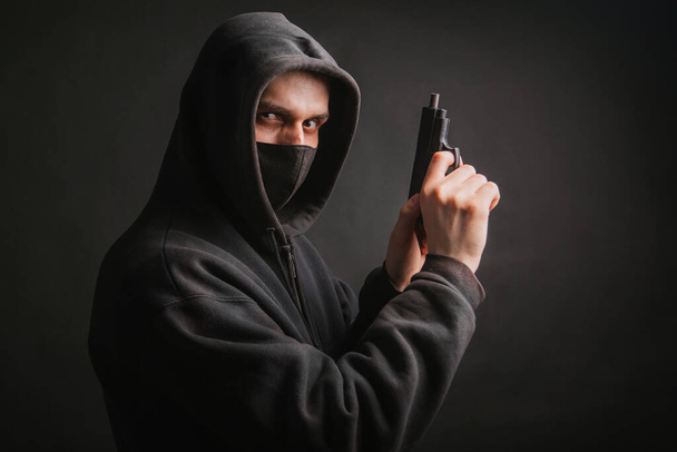 Een gangster in een capuchon, een masker, houdt een wapen in zijn handen op een donkere achtergrond. De bandiet mikt met een pistool. Foto in de studio. - Foto, afbeelding