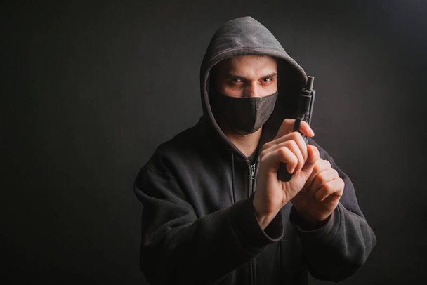 Egy csuklyás gengszter, egy maszk, fegyvert tart a kezében egy sötét háttérben. A bandita pisztollyal céloz. Fénykép a stúdióban. - Fotó, kép