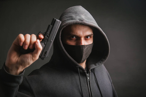 A gangster in a hood, a mask, holds a weapon in his hands on a dark background. The bandit aims with a pistol. Photo in the studio. - Foto, Imagem
