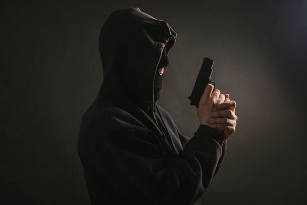 A gangster in a hood, a mask, holds a weapon in his hands on a dark background. The bandit aims with a pistol. Photo in the studio. - Photo, Image