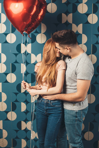 Charmant couple embrassant sur un mur bleu studio le jour de la Saint Valentin tenant des ballons à air rouge et s'embrassant - Photo, image