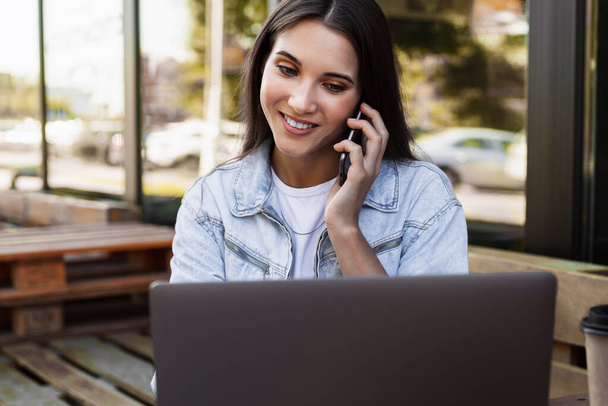 Belle femme d'affaires travaillant dans un café sur une terrasse ouverte, assise devant un ordinateur portable, parlant au téléphone.  - Photo, image