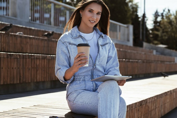 Millennial woman in denim clothes sits on park bench, holding a smart tablet in her hands. Woman uses tablet - Фото, зображення