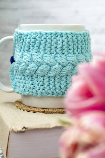 A white cup of tea in a knitted blue case stands on a book and pink flowers, tulips and gingerbread on a texture table. Spring concept. - Fotografie, Obrázek