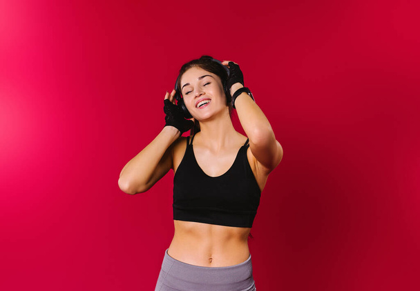 A cheerful girl with a beautiful physique with a smile listens to music in large headphones on a red background with empty side space. - Фото, изображение