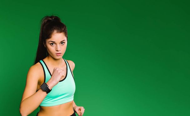 Young caucasian girl runner in light green sport top with a fitness watch on a green background looks at the camera while jogging. - Фото, изображение